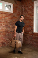 Man holding a tan duffel bag against a brick wall.