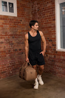 Man in black tank top and shorts holding a brown duffel bag against a brick wall.