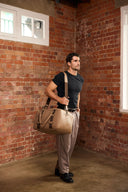 Man holding a beige leather bag against a brick wall.